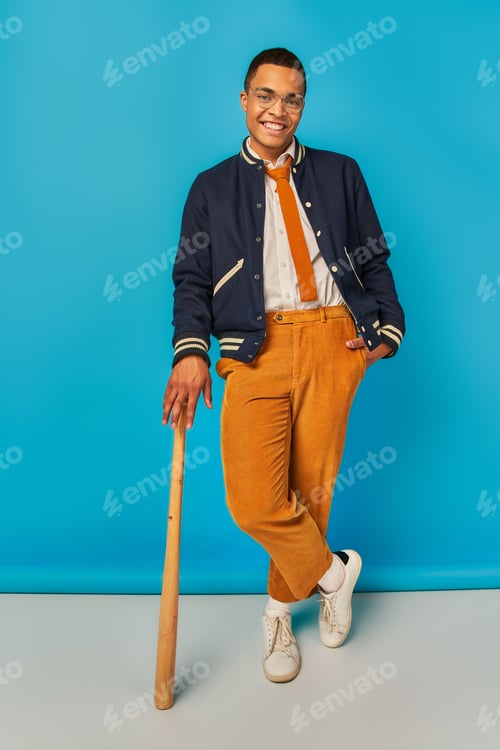 Preview: smiling african american student in jacket and orange pants posing with baseball bat on blue