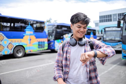 Preview: Happy Traveler Asian Male In Casual Style Smiling While Looking At Wristwatch Against Bus Station