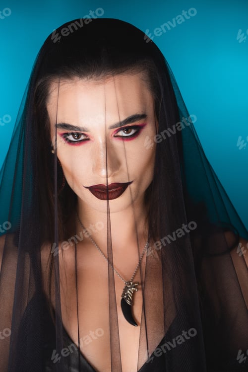 Preview: evil bride with dark makeup looking at camera through black veil on blue