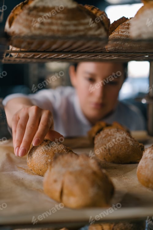 Preview: attractive female baker between shelves looking checking freshly baked bread very carefully