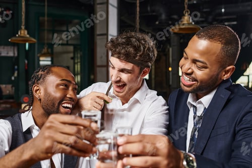 Preview: happy interracial friends toasting with glasses of tequila shots and laughing during bachelor party