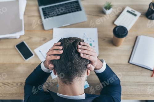 Preview: top view of depressed businessman covering head with hands while sitting at workplace near gadgets