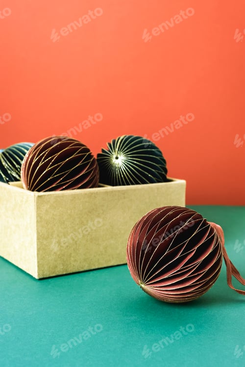 Preview: Christmas paper baubles in a cardboard box on green table with red background, copy space. Closeup
