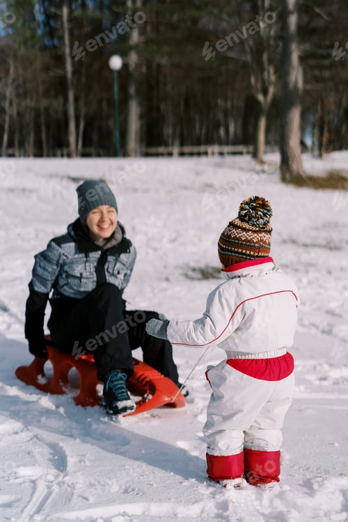 Preview: Small child rides a laughing mother on a sled on a snowy plain. Back view