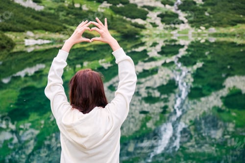 Preview: Young woman standing on the shore of the lake and showing a heart with her hands under head. Amazing