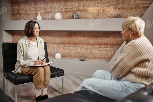Preview: middle aged psychologist in glasses smiling and talking to blonde woman during therapy session
