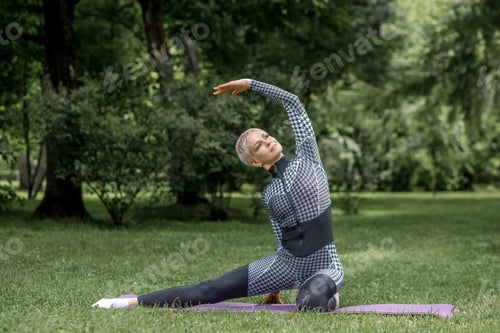 Preview: An elderly woman in sportswear does yoga on a mat outdoors.