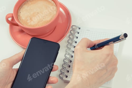 Preview: Hand of woman with mobile phone, coffee with milk and notepad for writing notes. White background