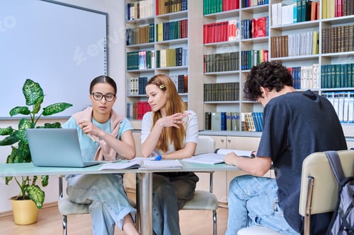 Preview: Group of students teenagers sitting together at desk in library preparing for tests exams