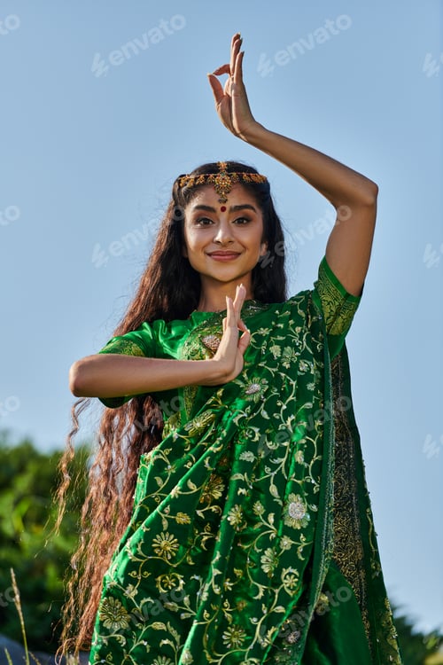 Preview: portrait of stylish young indian woman in sari and matha patti posing in park with blue sky