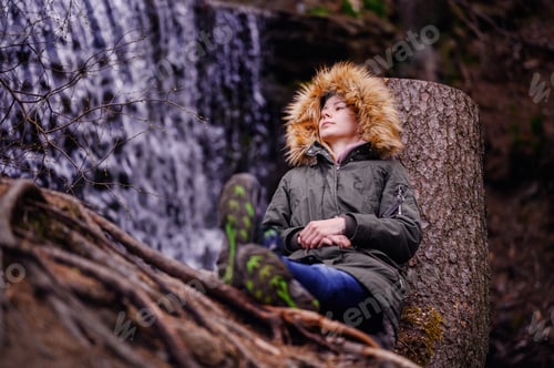 Preview: Young woman sits leaning on a stump in green parka with fur before waterfall alone in forest