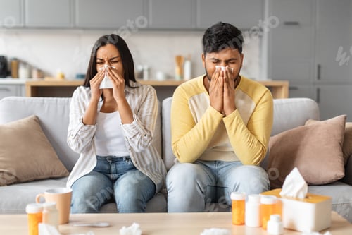 Preview: Sick indian couple sitting on sofa, blowing noses and suffering from colds