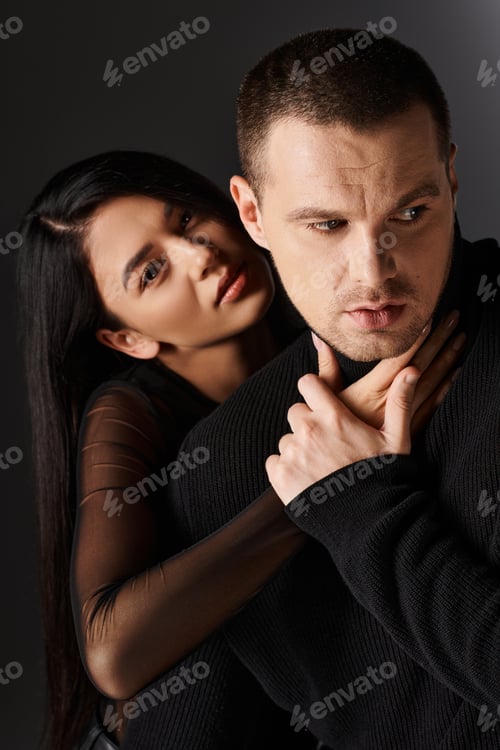 Preview: seductive asian woman embracing young man in black outfit and looking at camera on grey backdrop