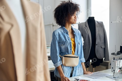 Preview: Dreamful African-American lady tailor stands at table in contemporary workshop