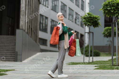 Preview: Muslim woman, wearing grey pants and green jacket, walking at the street with colorful shopping bags