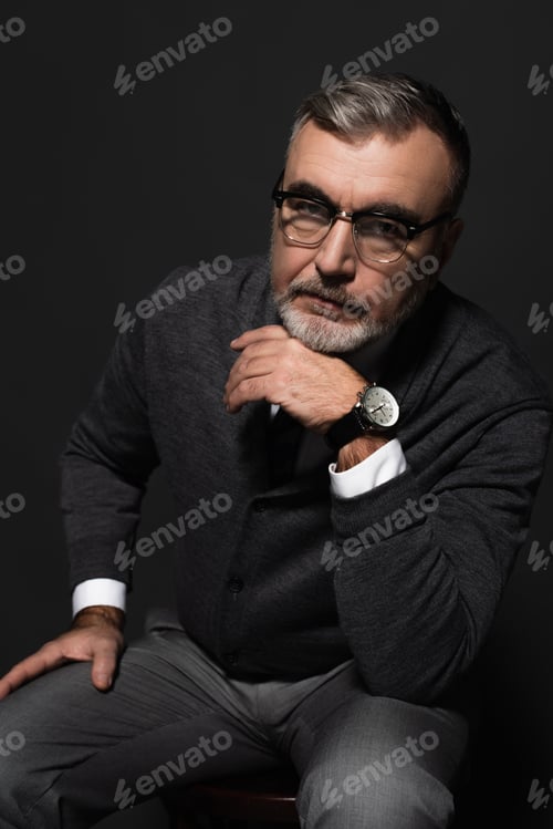 Preview: serious senior man in jumper, eyeglasses and wristwatch looking at camera while sitting on dark grey