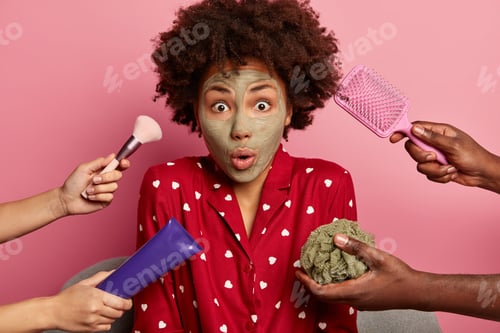 Preview: Emotive stupefied dark skinned woman in night clothes, applies clay mask, poses against pink backgro