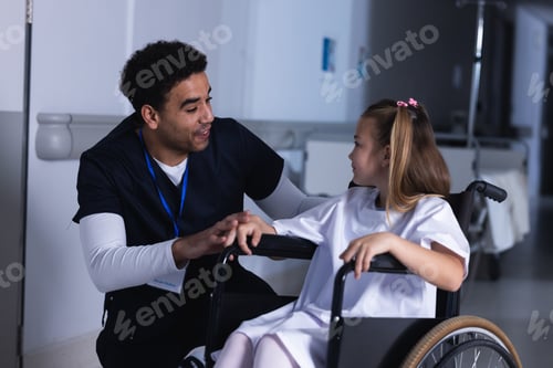 Preview: Diverse male doctor talking with girl in wheelchair in hospital corridor