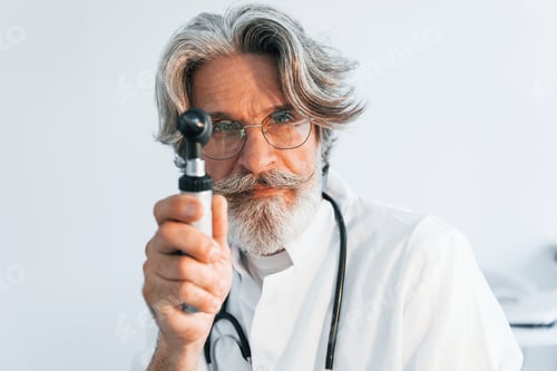 Preview: Portrait of senior male doctor with grey hair and beard in white coat is indoors