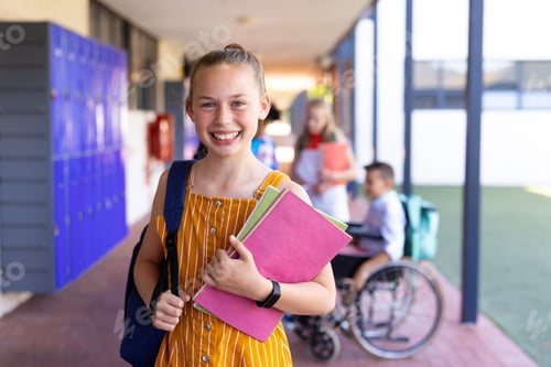Preview: Portrait of smiling caucasian of schoolgirl holding books in school corridor with copy space