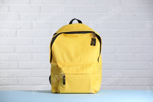 Preview: Yellow backpack on light blue table against white brick wall