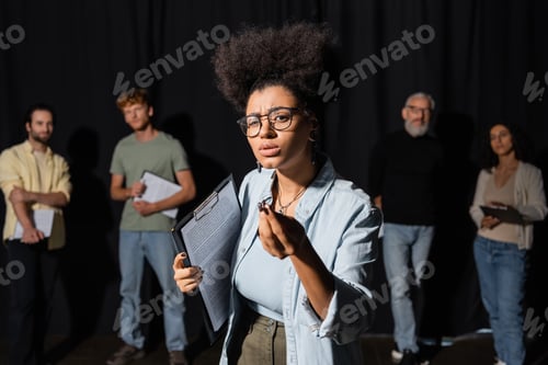 Preview: african american woman holding screenplay and gesturing during rehearsal near blurred actors and