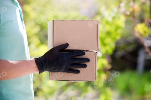 Preview: Delivery man holding cardboard boxes in medical rubber gloves at sunset on outdoor.