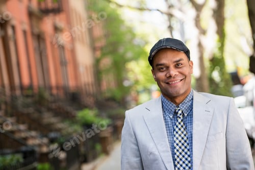 Preview: A man in a jacket and tie on a city street outside a terrace of brownstone houses.