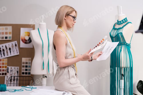 Preview: Concentrated designer with color swatch and sketch sits on desk in clothes atelier woman tailor
