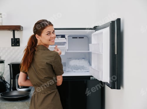 Preview: Woman smiling with teeth looking into camera in kitchen at home opened freezer empty with ice inside