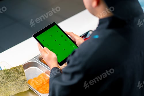Preview: Close-up from behind the sushi chef holding a tablet with a green screen open while making sushi