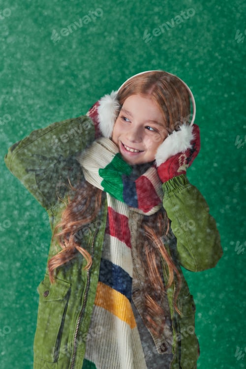 Preview: happy girl in ear muffs, striped scarf and winter attire standing under falling snow on turquoise
