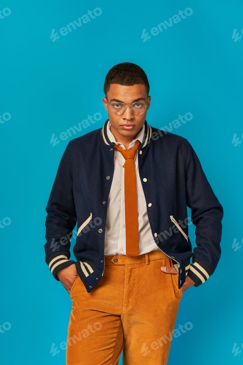 Preview: modern, trendy african american student in jacket, orange pants, with hands in pockets on blue