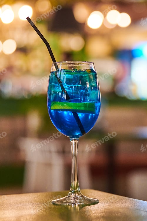 Preview: Blue Lagoon cocktail drink with lemon in glass with straw, night cafe light bokeh background