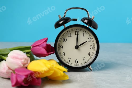 Preview: Spring time. Alarm clock and beautiful tulips on grey marble table against light blue background