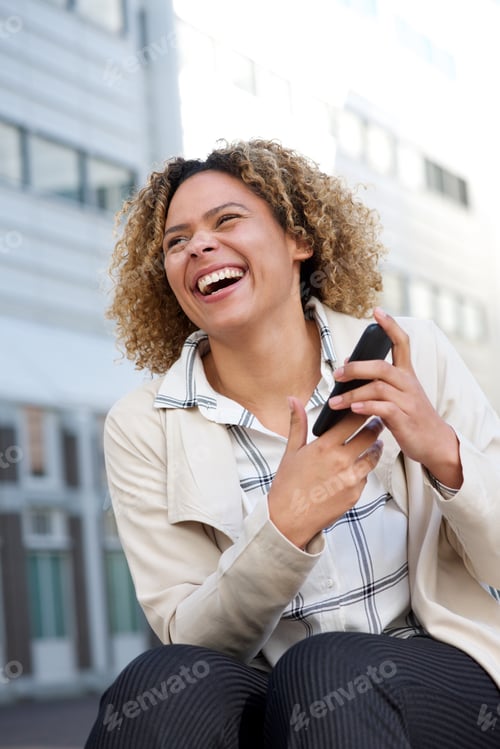 Preview: cheerful young african american woman laughing outside with cellphone