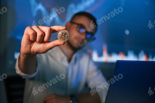 Preview: Holding physical bitcoin in hand. Stock broker trading online in the office with big screen