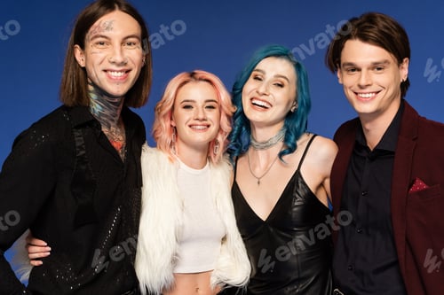 Preview: tattooed queer person looking at camera near trendy and cheerful nonbinary friends isolated on blue