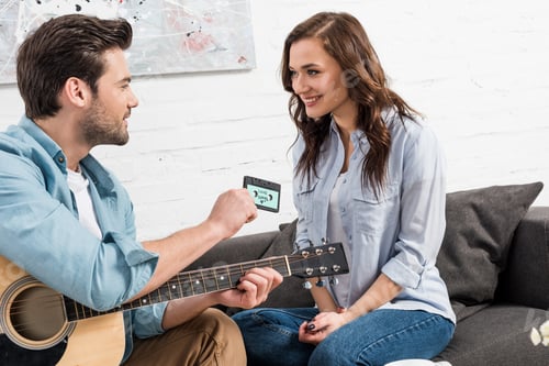 Preview: man with acoustic guitar sitting on couch and giving vintage audio cassette to beautiful woman at