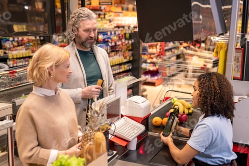 Preview: Young cashier scanning bottle of olive oil while looking at mature couple