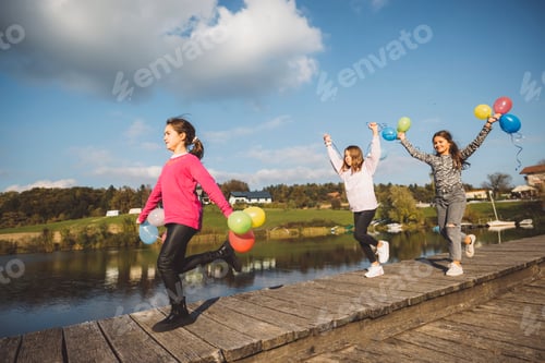 Preview: Three children joyfully running along a wooden pier by a lake, each holding colorful balloons. The s