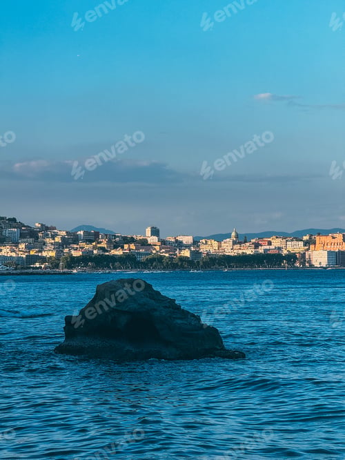 Preview: A large rock sits in the ocean near a city