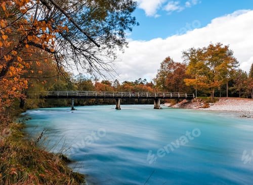 Preview: Long exposure image of footbridge across blue river surrounded with trees in autumn.