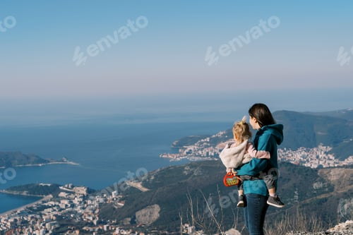 Preview: Mom with a little girl in her arms stands on a mountain and looks at the sea in the valley. Back