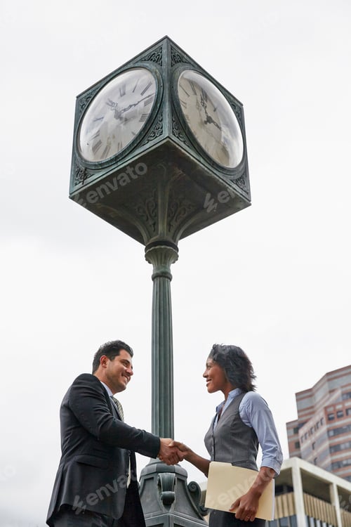Preview: Low angle view side view of business people shaking hands underneath clock, waist up