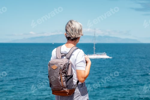 Preview: Rear view of senior woman with backpack standing and looking at sea and at the island at the horizon