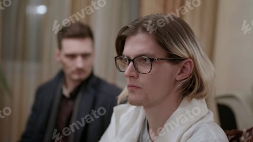 Preview: Two men sitting in a room in silence. Stock. Man with dyed long blond hair wearing glasses.