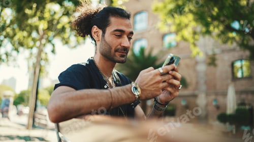 Preview: Attractive italian guy using mobile phone sitting on bench at old buildings background