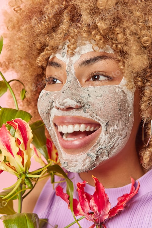 Preview: Headshot of cheerful woman with natural curly hair smiles broadly applies beauty mask on face holds