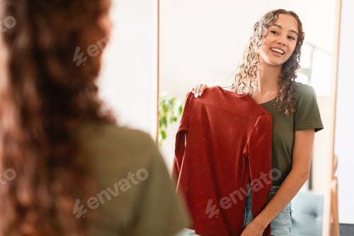 Preview: Happy caucasian teenager girl holding longsleeve standing near mirror indoors